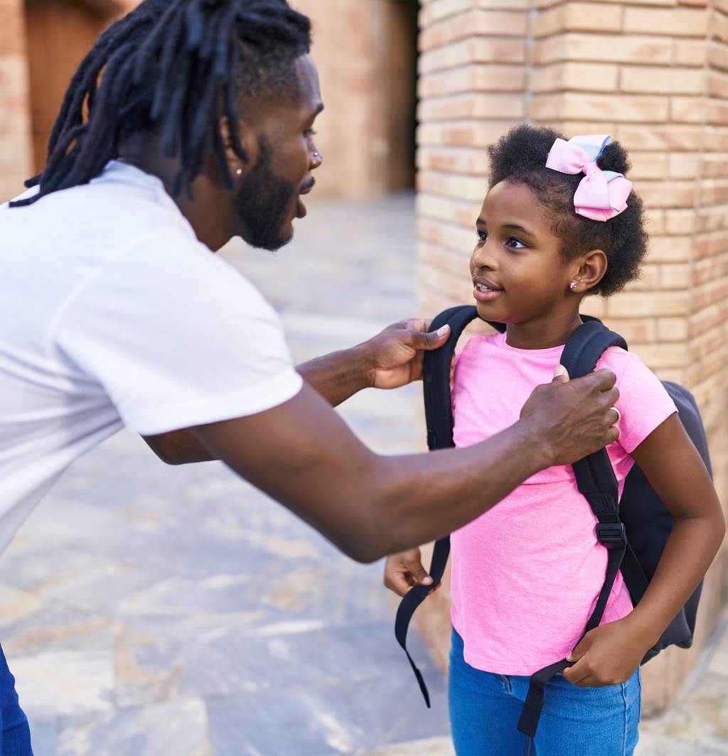 A father helps his school-age daughter put her backpack on.