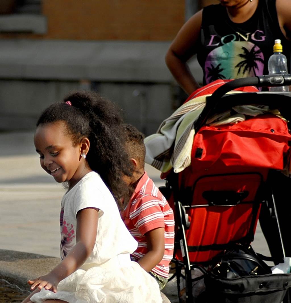 A mother with a stroller stands behind two young smiling children on a bench.