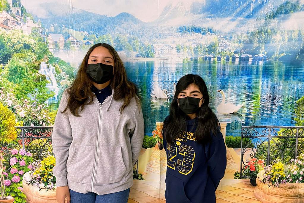 A photo of Itzel and Guadalupe in front of a tapestry in their school