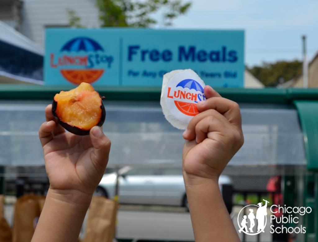 Two child-sized hands. One hand is holding a half eaten plum and the other is holding a sticker that says "LunchStop."