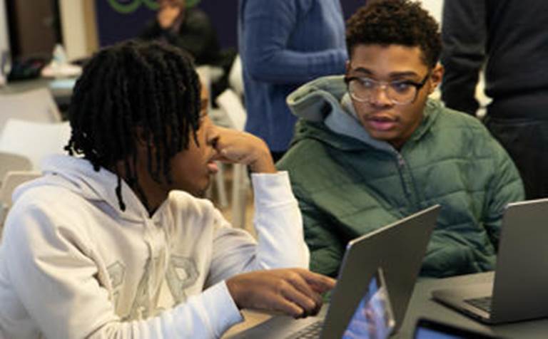 Two young male students sit side-by-side, collaborating and looking intently at a laptop screen in a classroom.