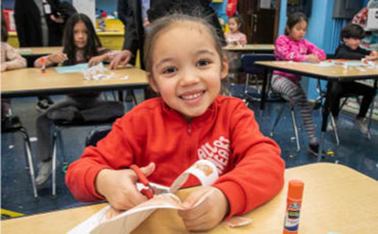 A young girl in a bright red sweater smiles enthusiastically for the camera while sitting at her desk and cutting paper for an arts and crafts project