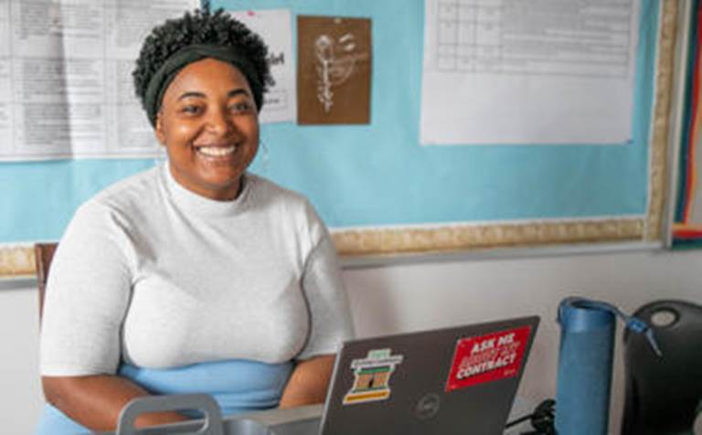 A smiling female teacher with short hair sits at her desk in front of a classroom bulletin board, looking warmly toward the camera.