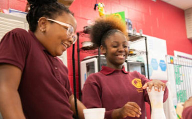 Two smiling young female students in school uniforms work together on a hands-on activity in their classroom.